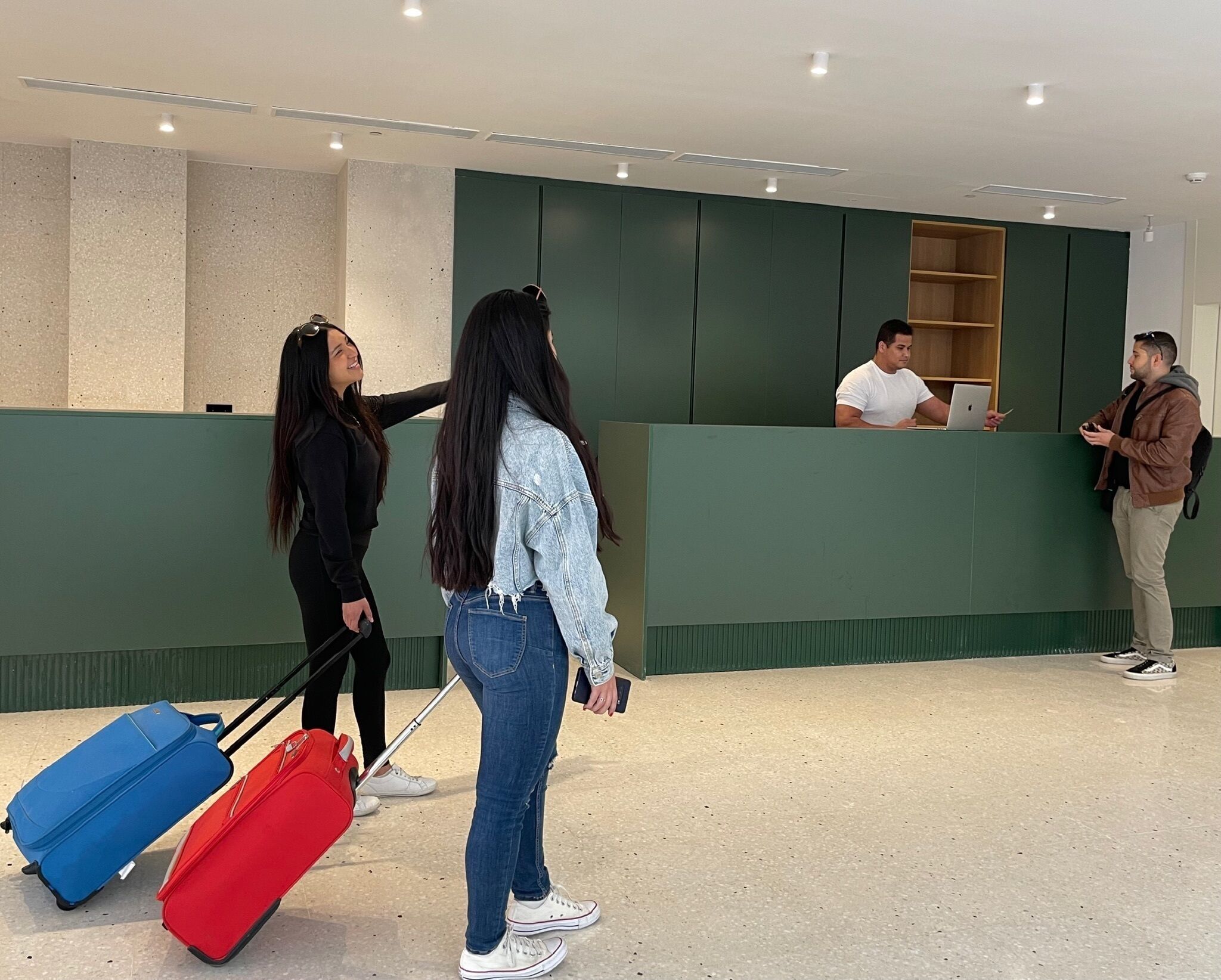 Un área de recepción con mostrador, un hombre trabajando y dos personas con equipaje.
