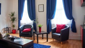 Un salon avec deux fauteuils en cuir noir, une petite table et un tapis bleu. On y trouve deux plantes en pot et un tableau encadré accroché au mur.
