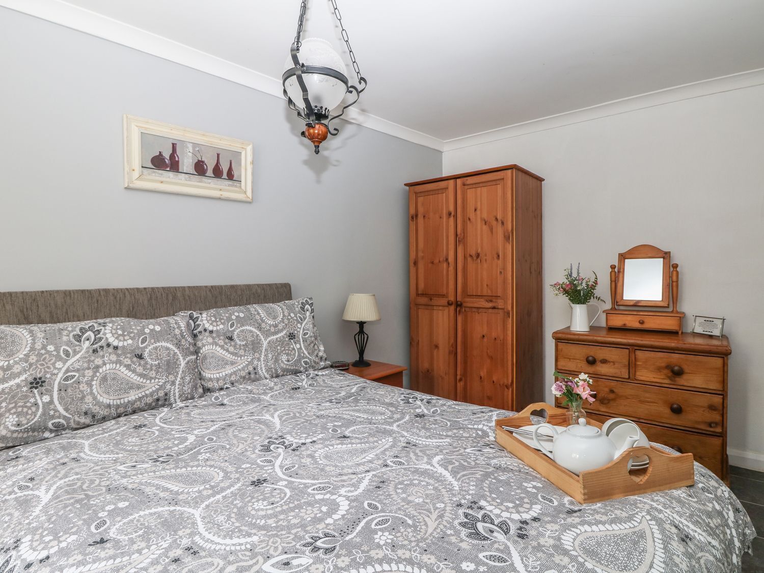 A bedroom with a bed, a wooden wardrobe, a nightstand, a lamp, and a mirror.