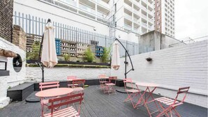 Une terrasse extérieure avec des chaises et des tables en métal rouge, des murs en briques blanches et une vue sur le paysage urbain.