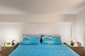 A bed with a blue ocean-themed bedspread, two pillows, a nightstand with a lamp, and a small decorative item on the nightstand.