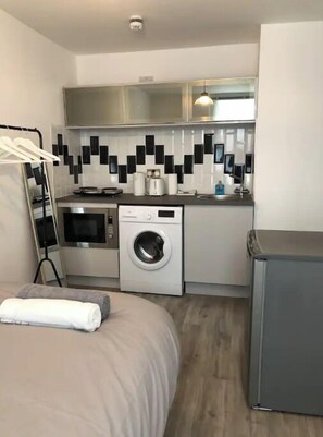 Une chambre d’hôtel compacte comprenant un lit, un lave-linge, un four à micro-ondes, une petite kitchenette et un meuble gris.