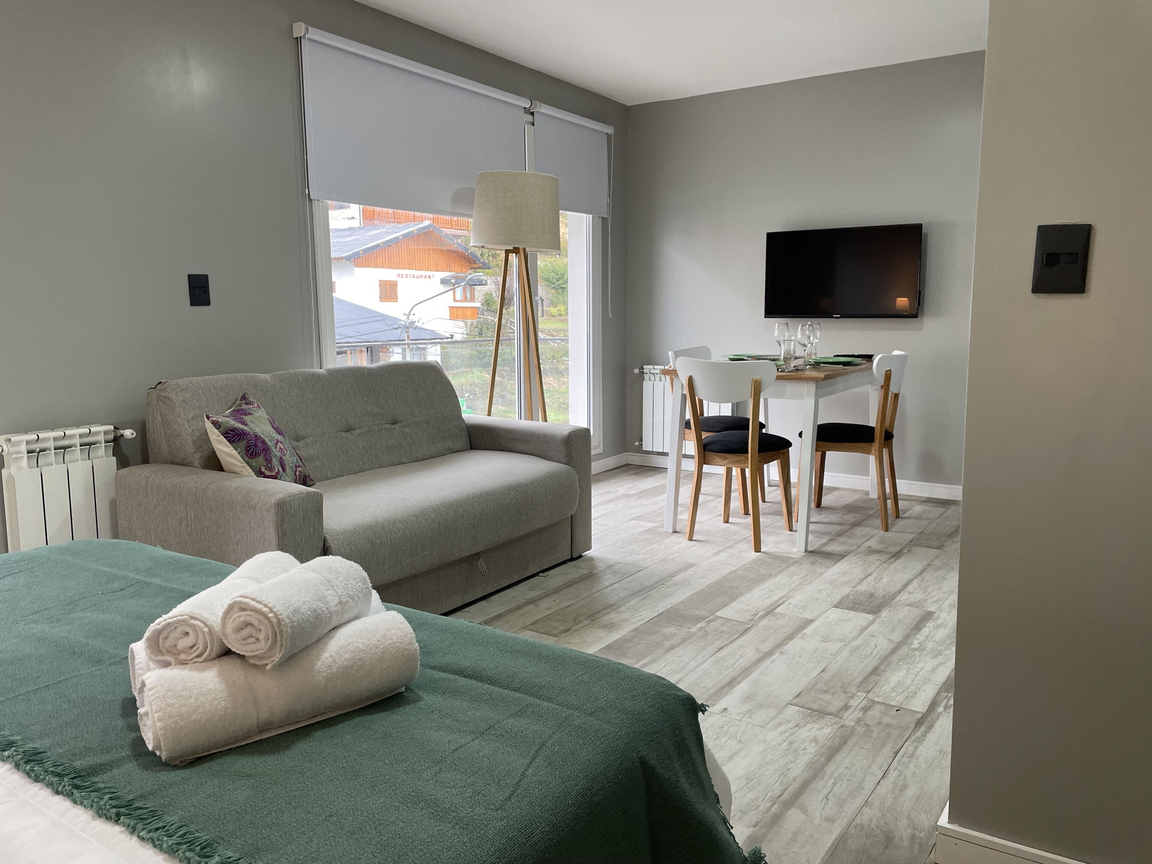 Une chambre d’hôtel moderne équipée d’un lit, d’un canapé, d’une table à manger avec des chaises, d’une télévision et d’une fenêtre donnant sur les bâtiments environnants.