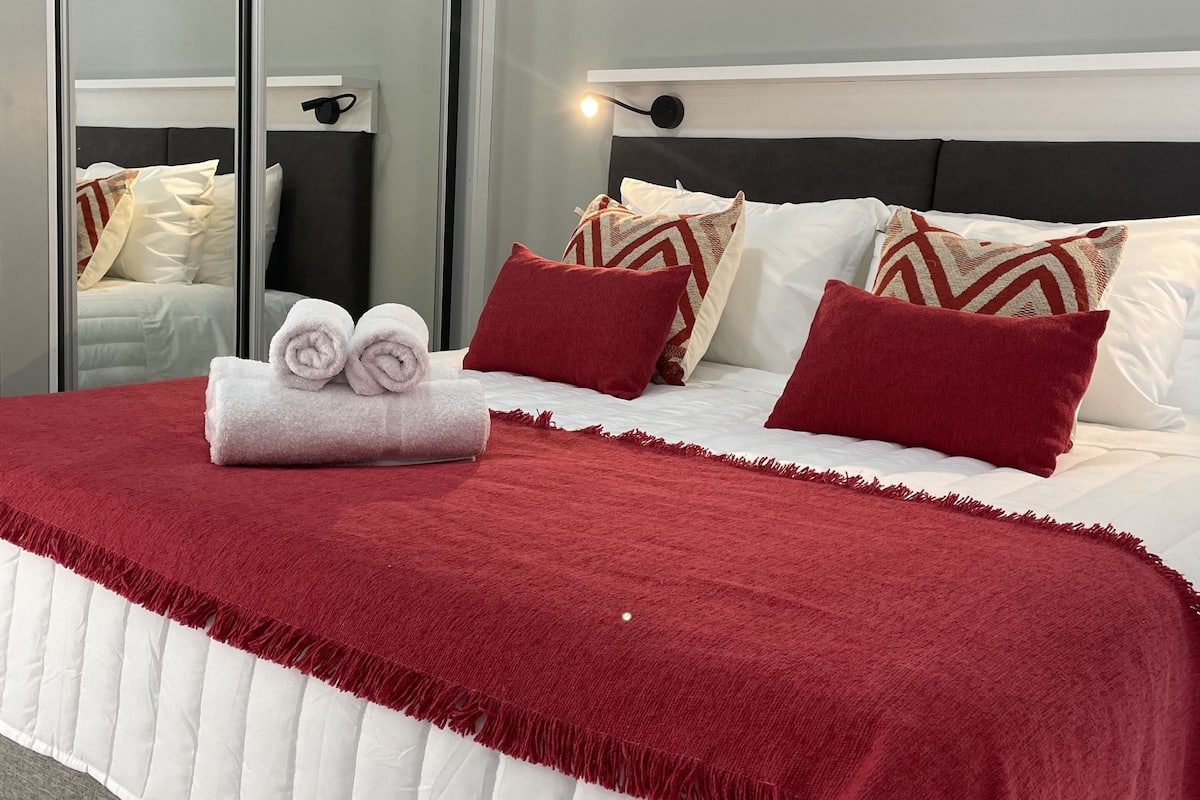 Un lit bien fait, avec une couverture rouge et des oreillers, une lampe de chevet et un miroir qui reflète la pièce.