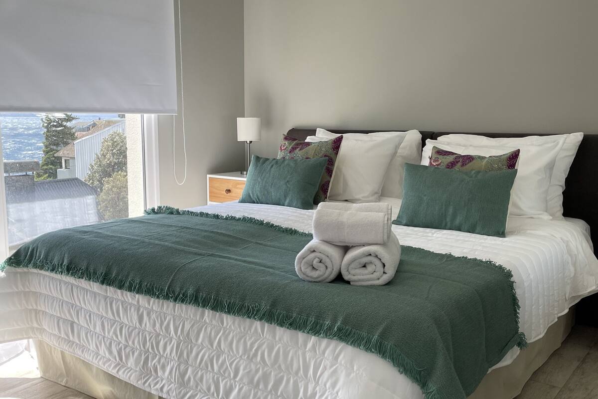 Une chambre à coucher avec un lit, des oreillers, une table de chevet avec une lampe, et une vue sur l’extérieur.