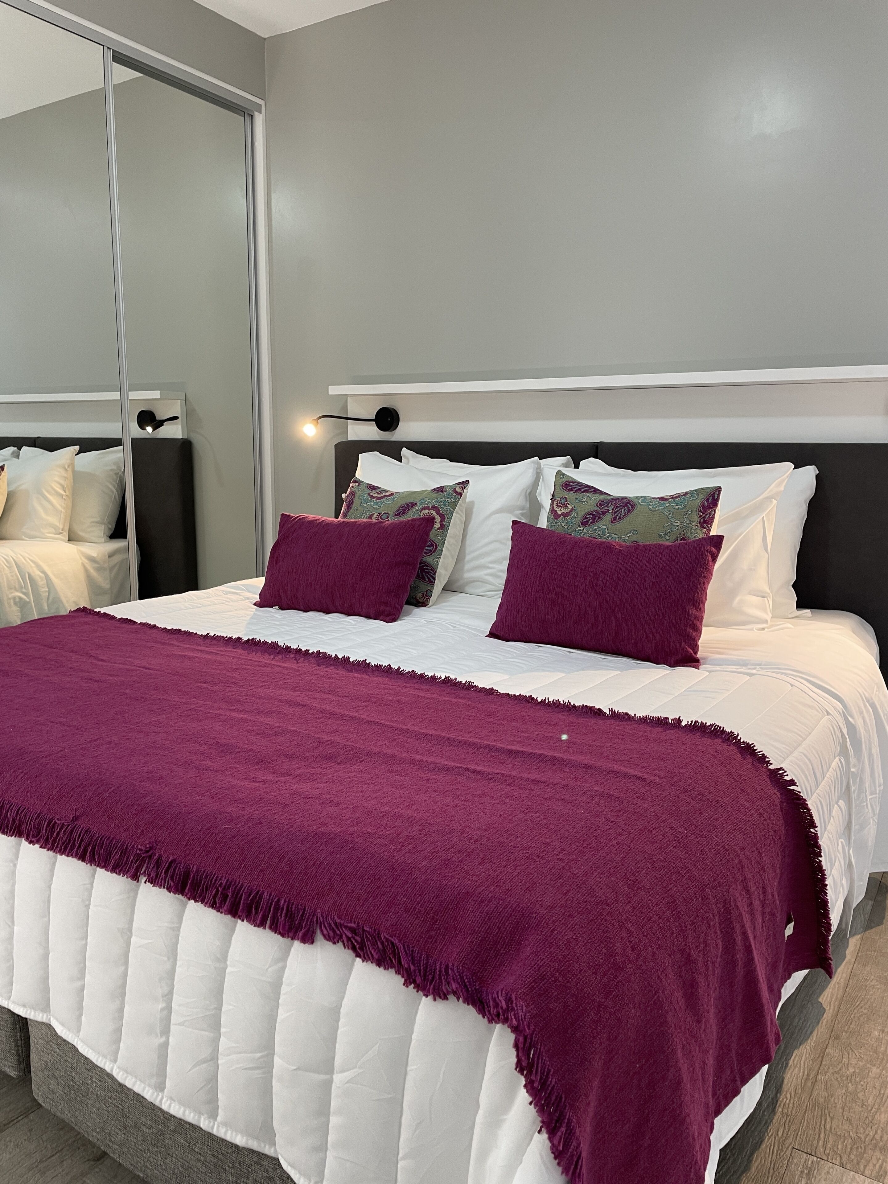 Una cama bien hecha con colcha blanca y morada, varios almohadones y un cabecero.