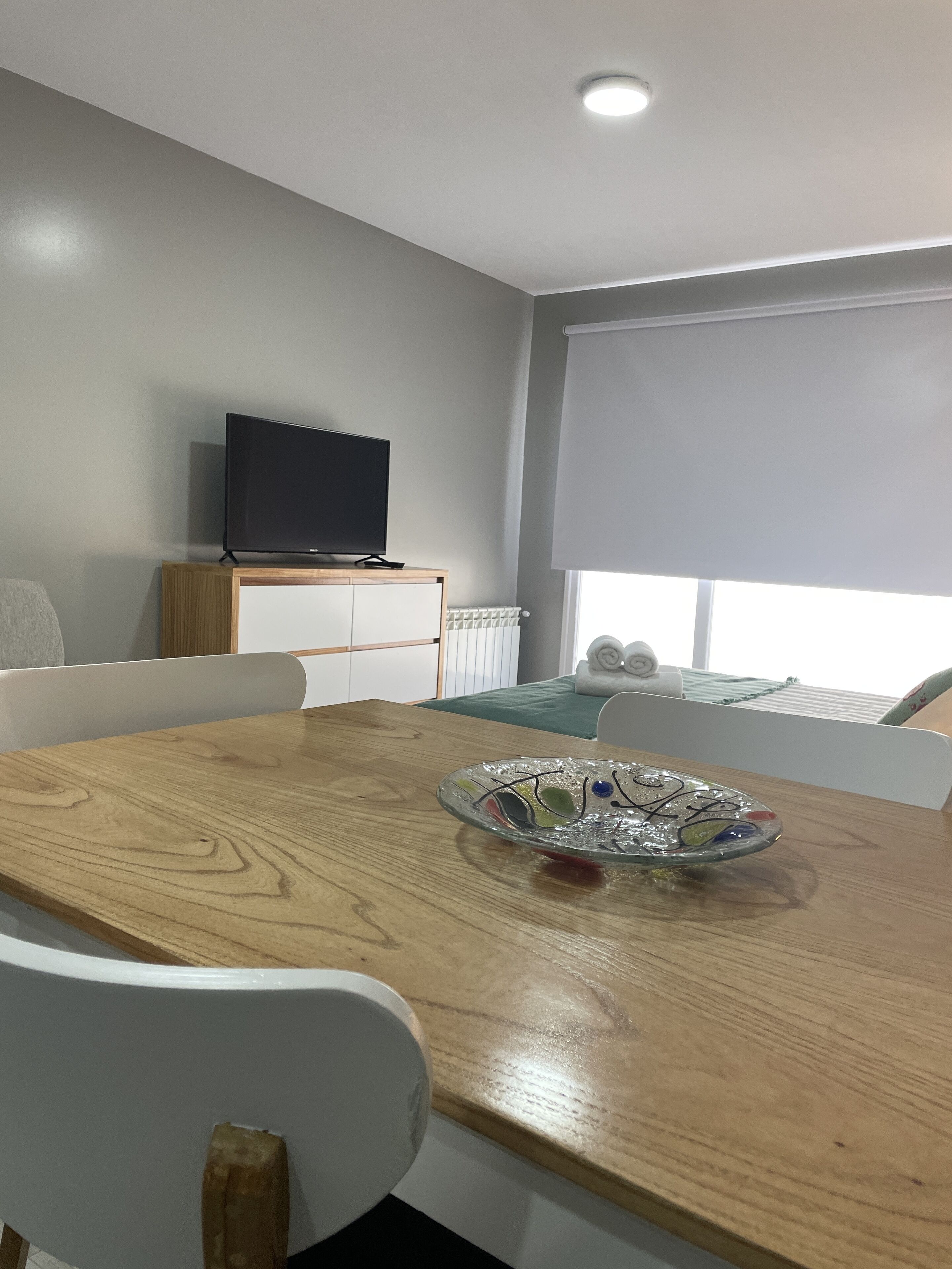 Una habitación moderna con una mesa de comedor de madera, sillas blancas, un televisor de pantalla plana sobre un mueble de madera, un ventanal grande con persiana blanca y una cama con ropa de cama verde.