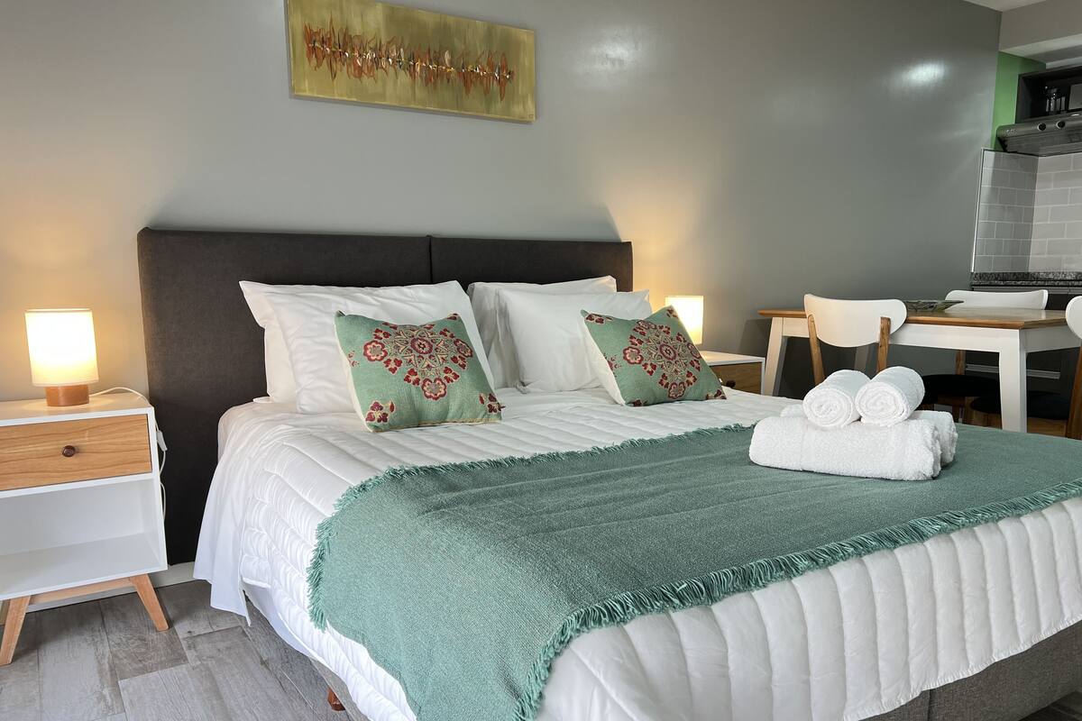 Un lit bien fait, agrémenté d’un plaid vert, de draps blancs et de coussins décoratifs. On y trouve une petite table de chevet avec une lampe et une table de nuit en bois.