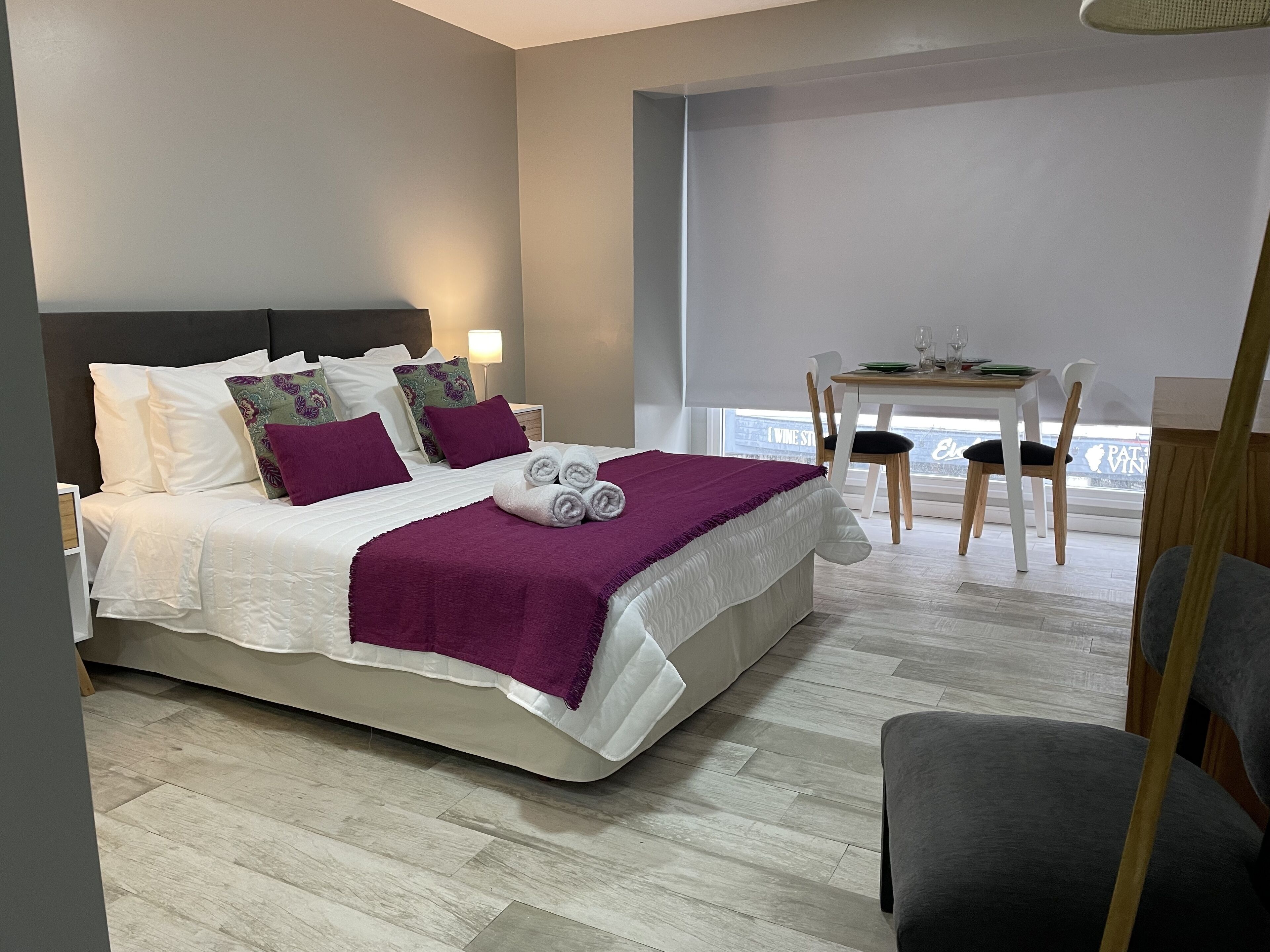 A modern hotel room with a neatly made bed, a dining table with chairs, and a view of the city through large windows.