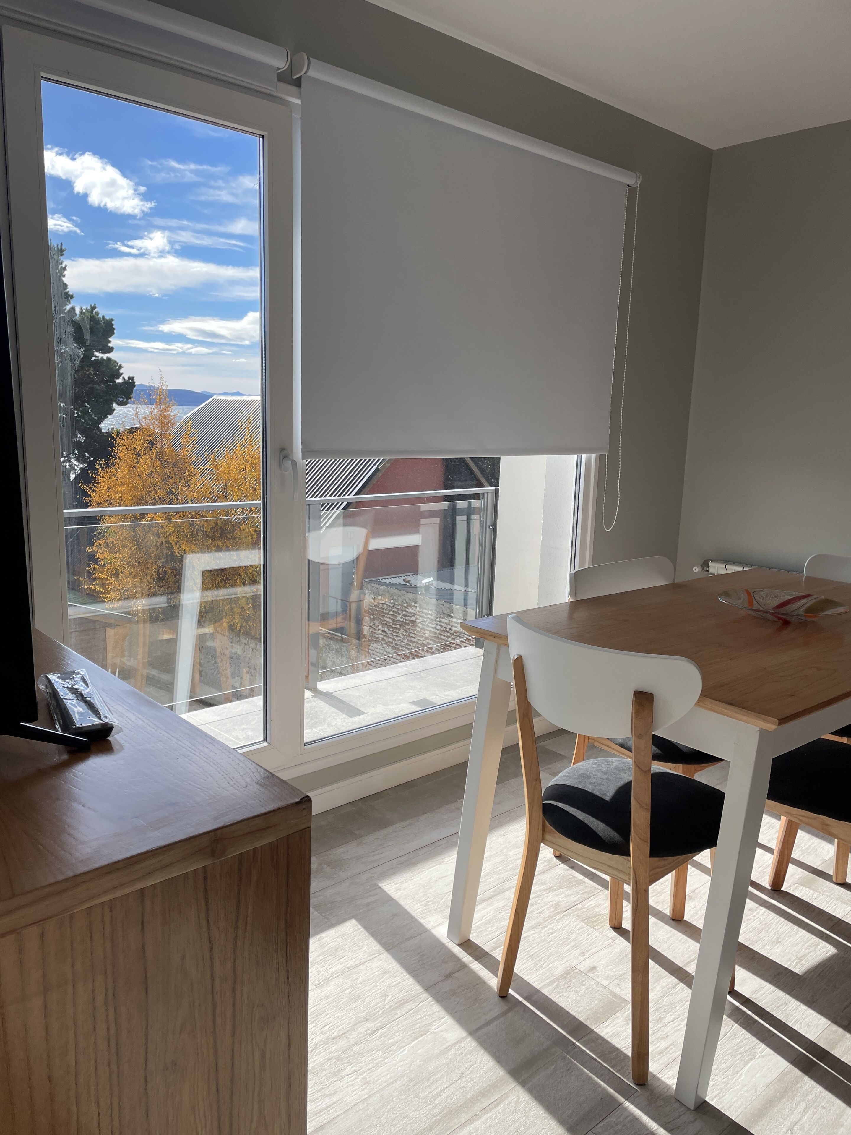 A modern room with a wooden table and chairs, a sliding glass door to a balcony, and a view of autumn trees and a distant mountain.