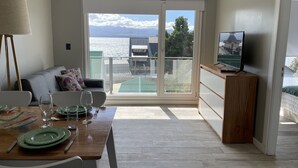 Een moderne woonkamer met een eethoek, een bank, een tv op een houten dressoir, en een balkon met uitzicht op zee en gebouwen.