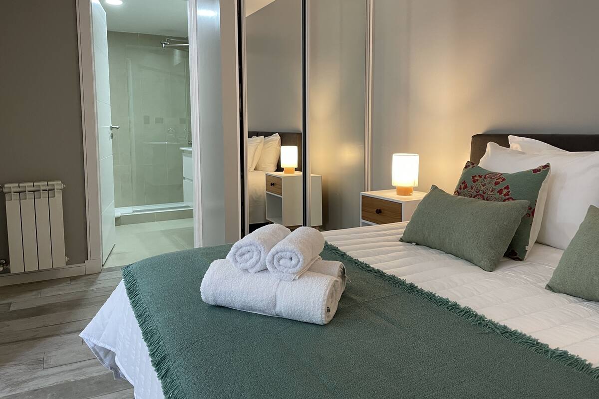 Une chambre d’hôtel avec un lit, une table de chevet, un miroir et une salle de bain visible à travers une porte ouverte.