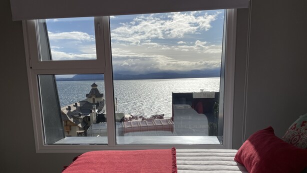 Een slaapkamer met een groot raam dat uitkijkt op een wateroppervlak en gebouwen, een bed met een rode deken en witte handdoeken, en een rood kussen.