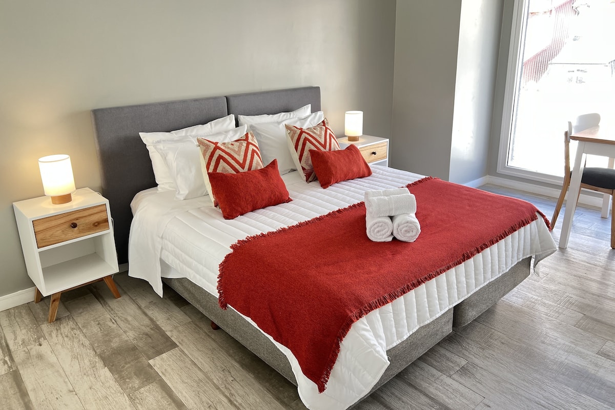 Un lit bien fait, avec du linge de lit blanc et des touches de rouge, une table de chevet avec une lampe, et une chaise près de la fenêtre.