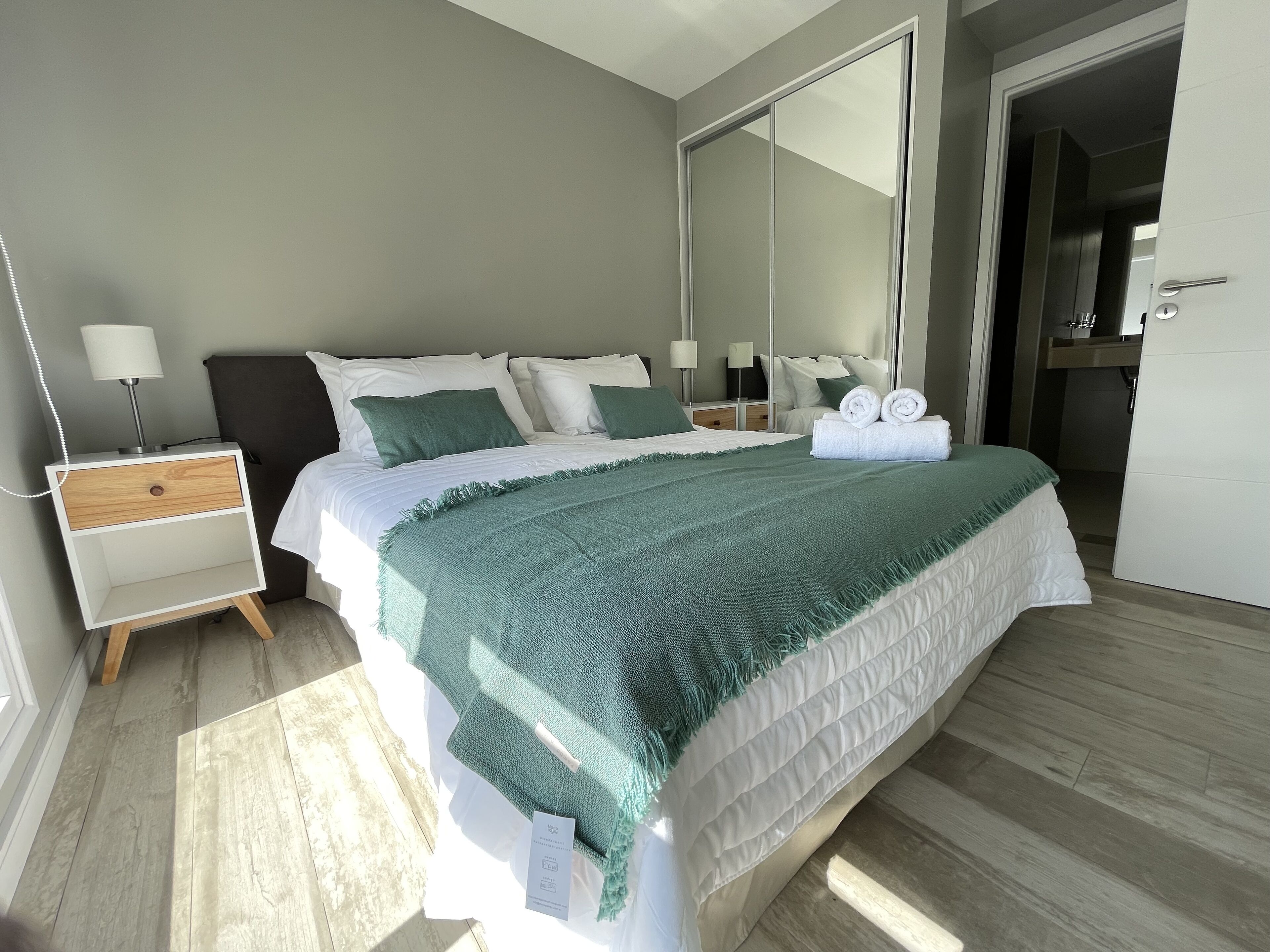 A neatly made bed with a green blanket, a bedside table with a lamp, and a large mirror on the wall.