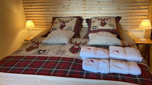 Un lit recouvert d’une couverture à carreaux rouges et noirs, deux oreillers ornés de motifs de rennes et quatre serviettes pliées.