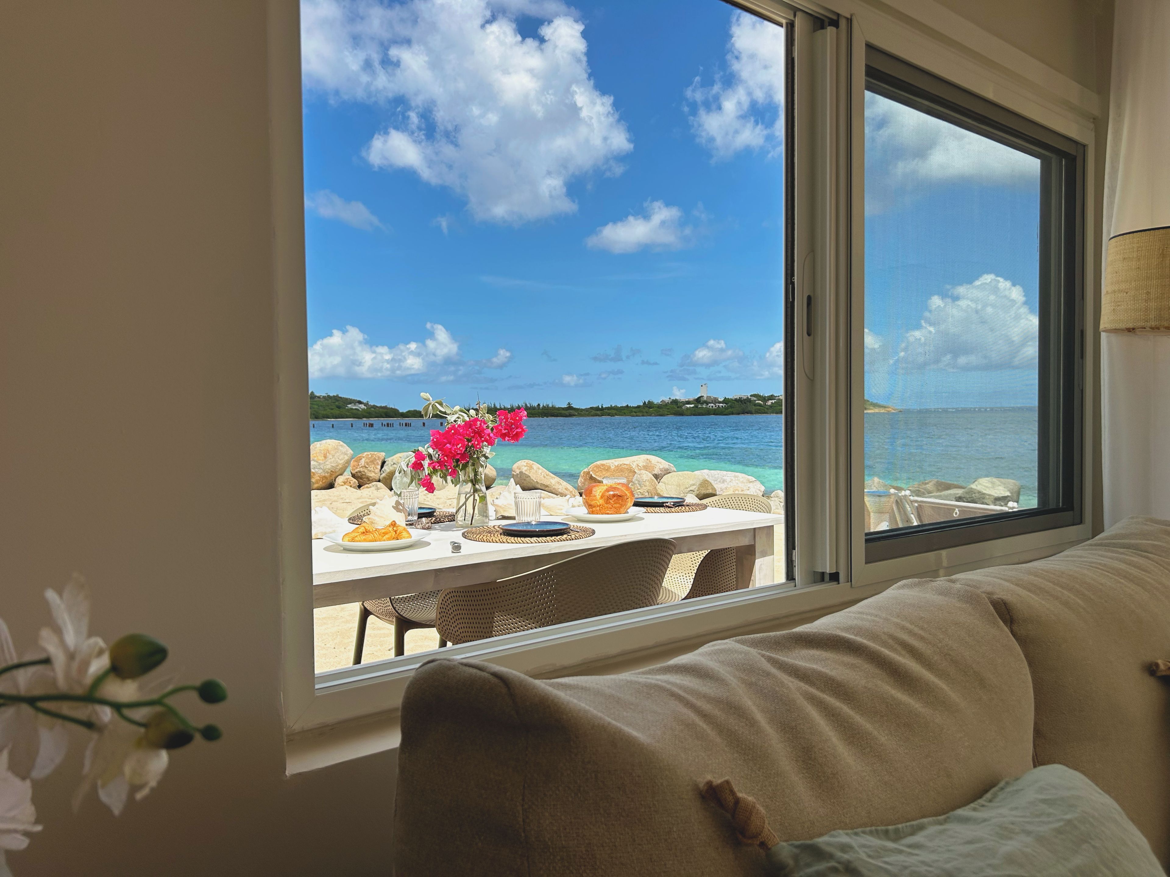 A room with a view of a beach and sea, a dining table set with plates and a vase of flowers.