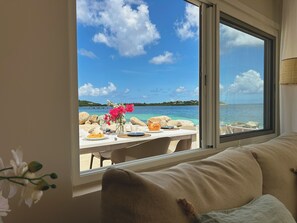 A room with a view of a beach and sea, a dining table set with plates and a vase of flowers.