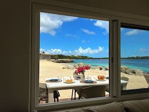Una mesa de comedor con platos, un jarrón con flores y vistas a una playa y al agua a través de una ventana.