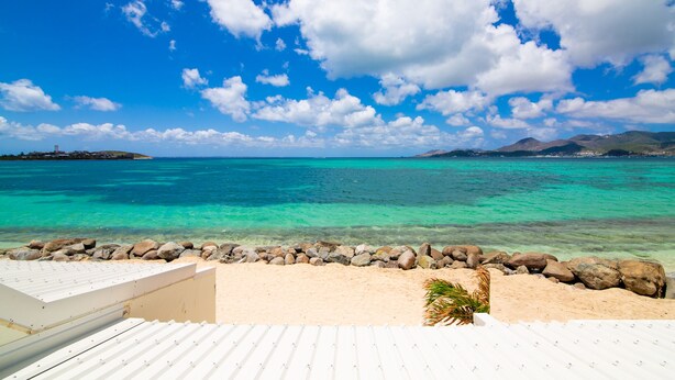 A beach view with clear turquoise water, a rocky shoreline, and a white building with a sloped roof.