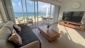 A modern living room with a large TV, a sofa with patterned cushions, a coffee table, and a view of the beach and ocean through large windows.
