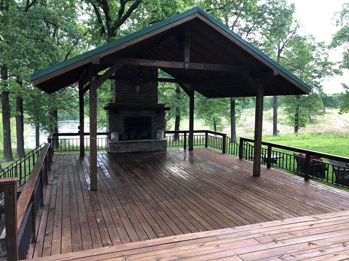 A covered wooden deck with a stone fireplace, surrounded by a metal railing and trees.