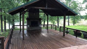 A covered wooden deck with a stone fireplace, surrounded by a metal railing and trees.