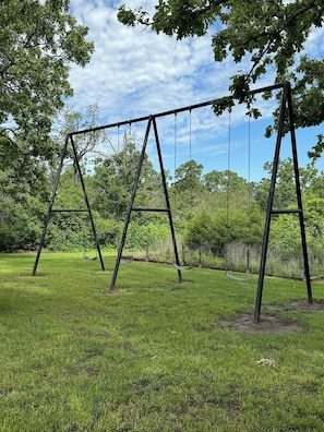 Un columpio en un parque con césped y árboles al fondo.