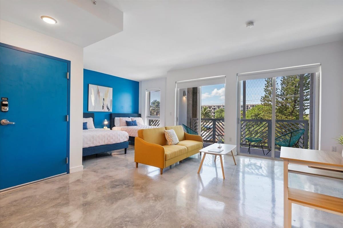 A modern hotel room with a blue accent wall, two beds, a yellow sofa, a balcony with outdoor seating, and a large window offering a view of the outdoors.