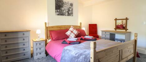 A bedroom with a wooden bed, a nightstand with a lamp, a desk with a chair, a red chair, a mirror, and a wall-mounted picture.