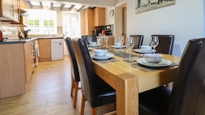 A dining area with a wooden table set for six, surrounded by black leather chairs. A kitchen with wooden cabinets and a dishwasher is visible in the background. - Tan Y Capel (Llanbrynmair)