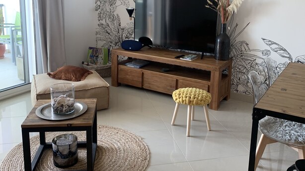 A modern living room with a flat-screen TV, a wooden entertainment unit, a coffee table, a yellow stool, and a dining table. - Cosy T3 100m from the beach in a recent residence. (Fleury D'aude)