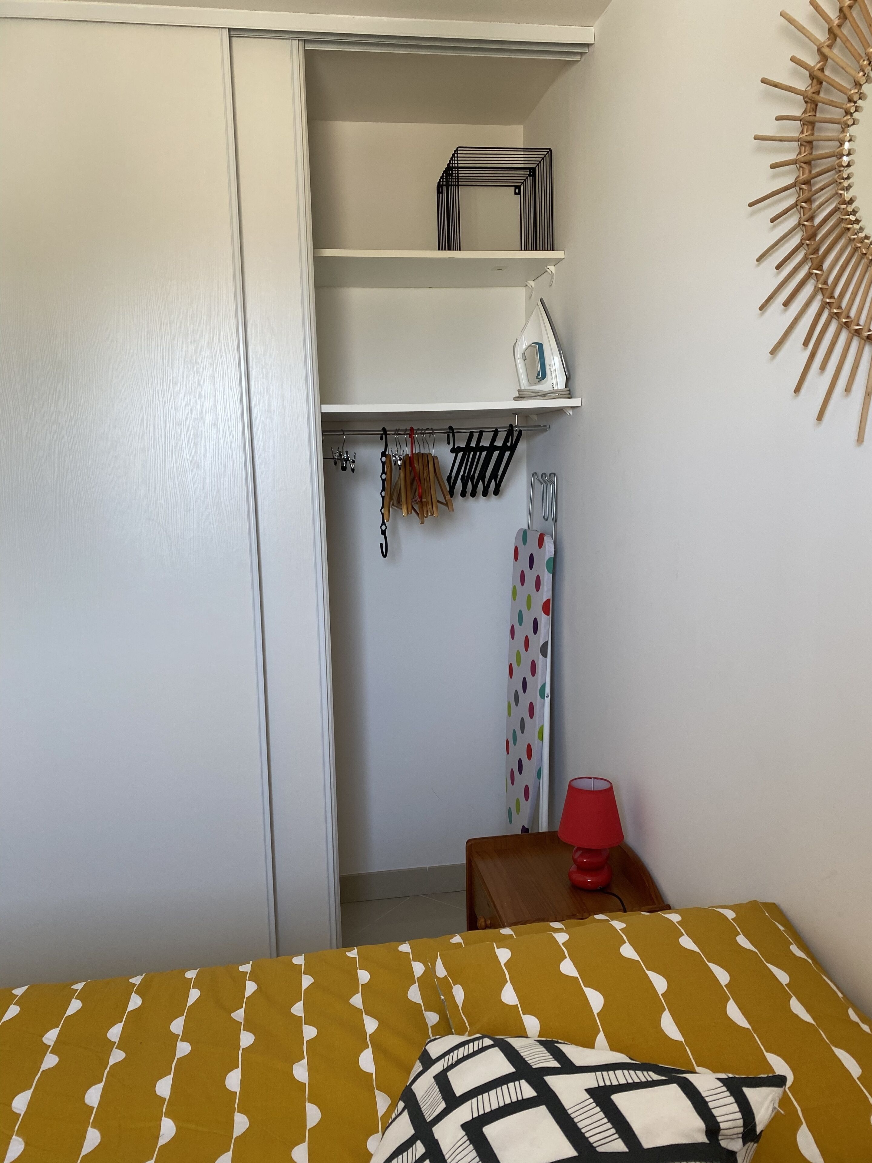 Een slaapkamer met een bed, een kledingkast, een plank met een strijkijzer en een wandlamp.