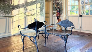 Quarto com lustre, duas cadeiras de metal, uma estátua e vista através de grandes janelas.