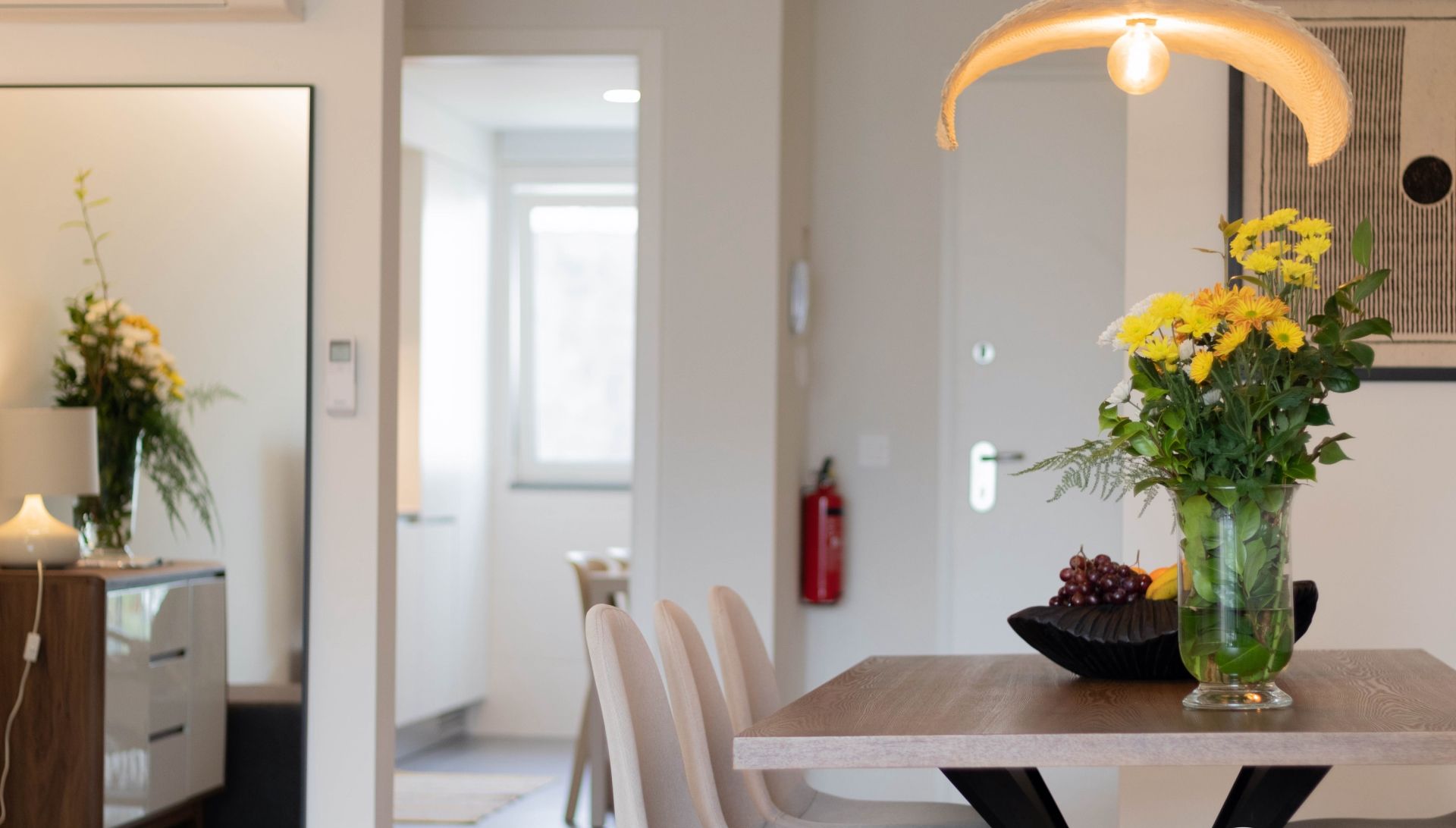 A modern dining area with a wooden table, white chairs, and a vase of yellow flowers.