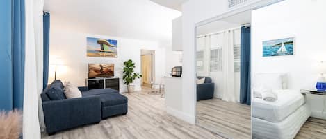 A modern living room with a sofa, a dining area, and a mirrored wall reflecting a bed.