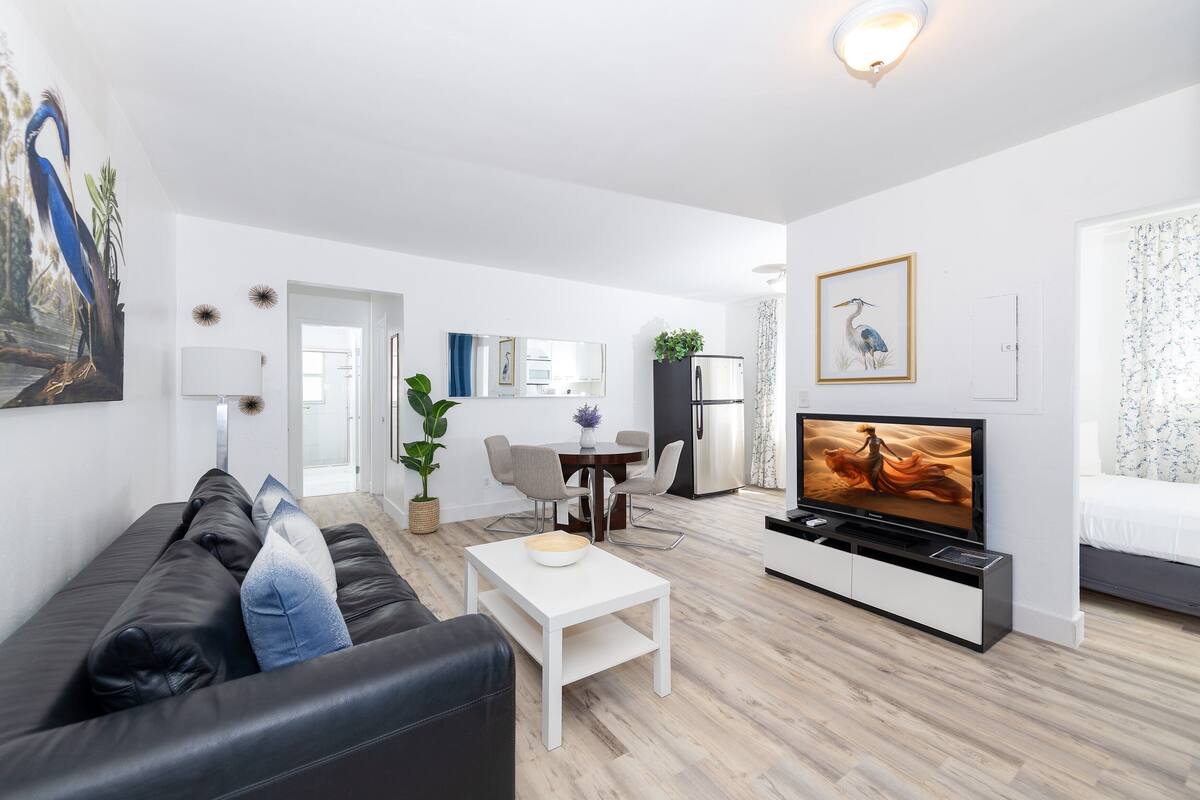 En moderne stue med en sort lædersofa, et hvidt sofabord, et fjernsyn og et spiseområde med bord og stole.
