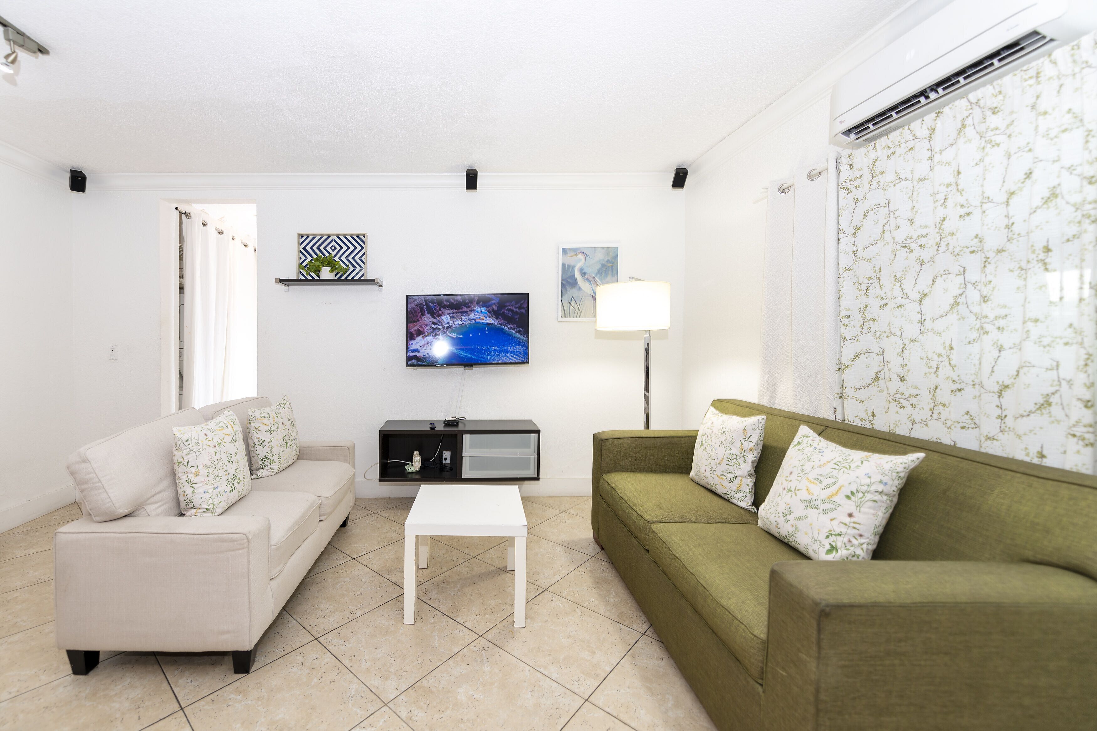 A living room with a beige sofa, a green sofa, a white coffee table, a flat-screen TV, a floor lamp, and a wall-mounted shelf with a decorative item.