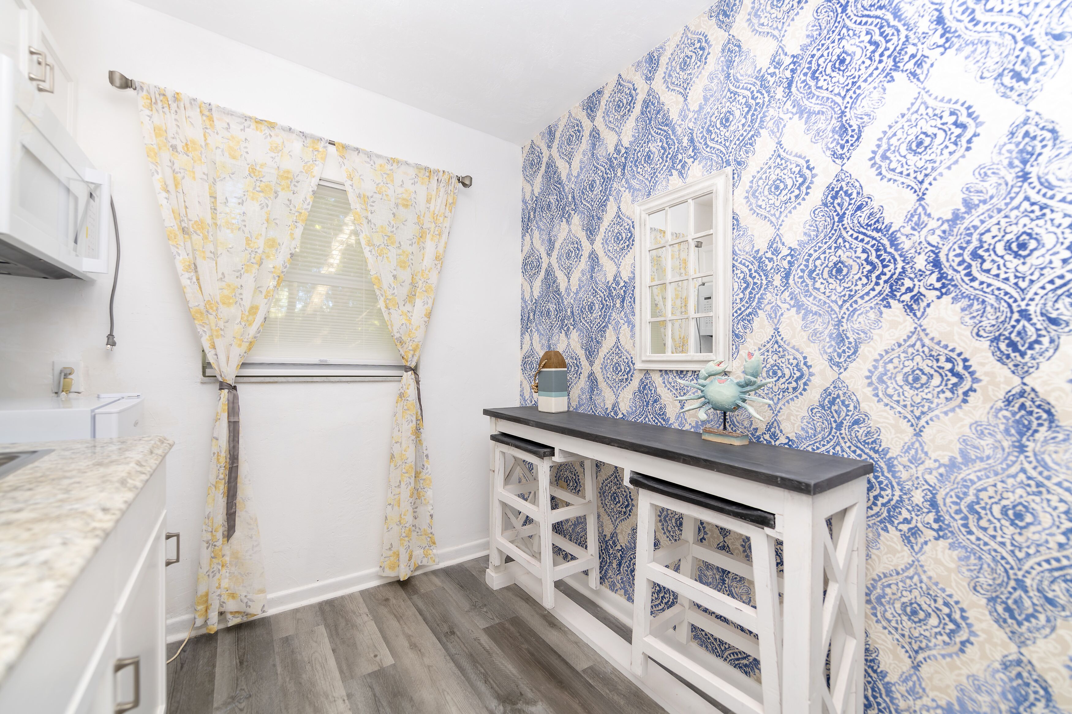 Una cocina con mostrador blanco, un taburete blanco y una ventana con papel tapiz estampado en azul.