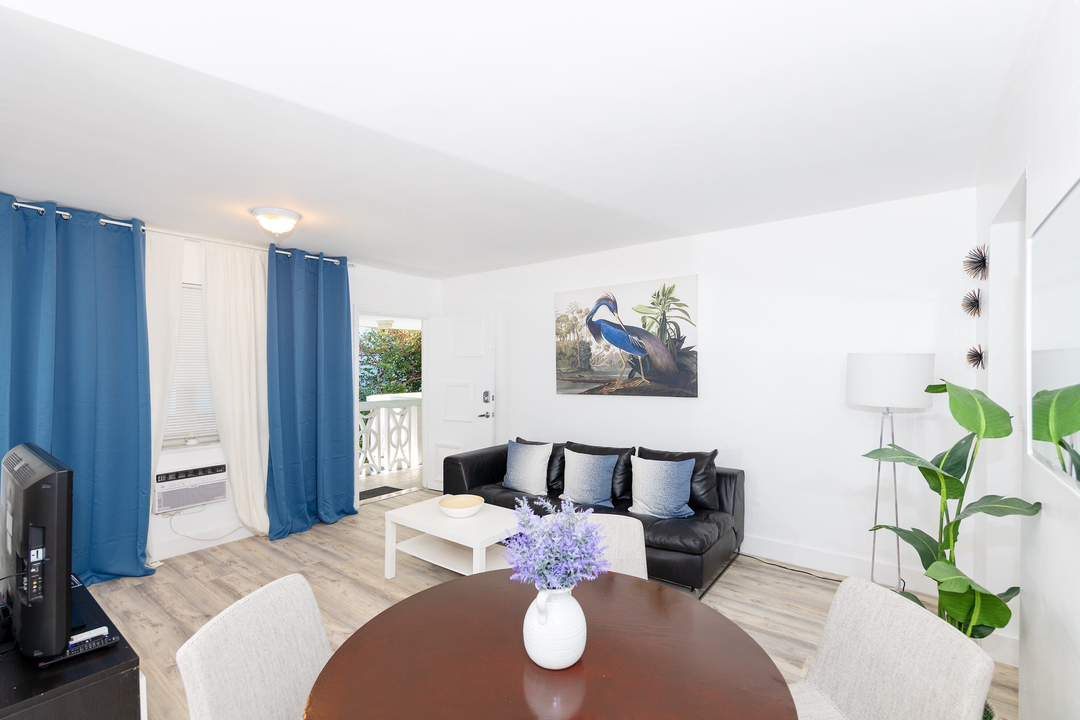 A modern living room with a black leather sofa, a white coffee table, and a dining table with a vase of flowers.