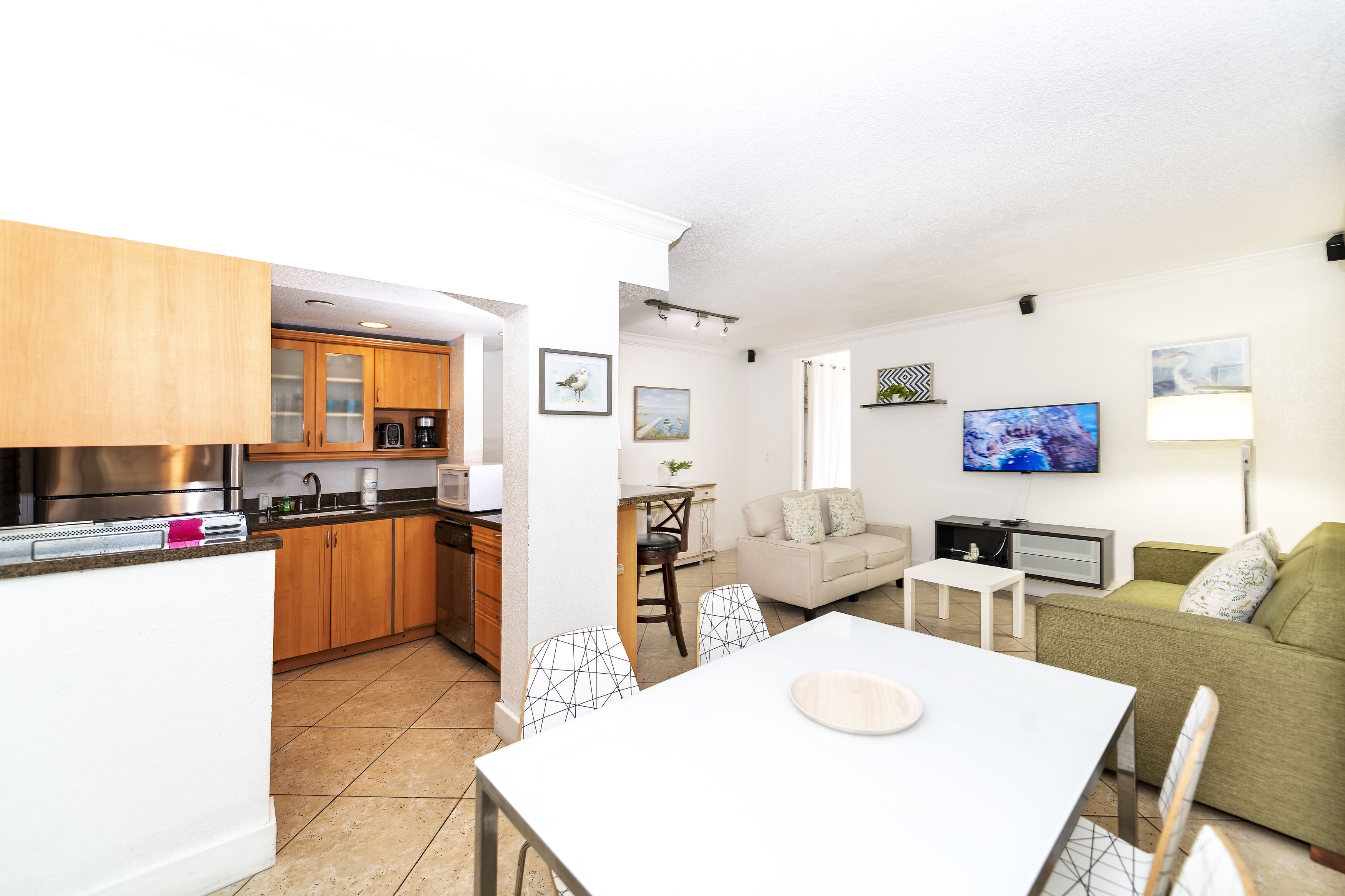 A modern kitchen with wooden cabinets, a dining area with a white table and chairs, a living room with a green sofa and a TV, and a small dining table with a lamp.