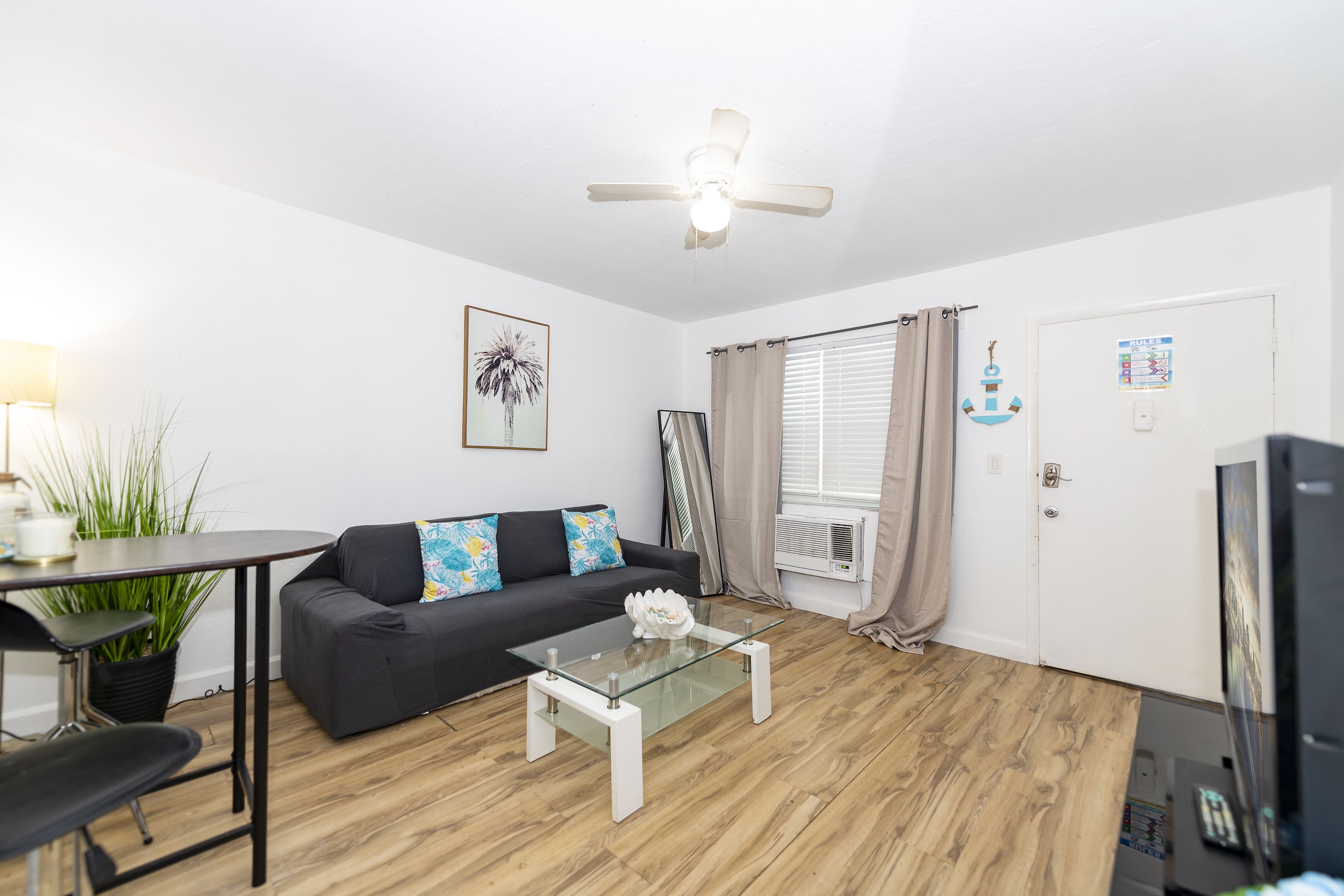 Ein modernes Wohnzimmer mit einem Glastisch, einer schwarzen Couch mit bunten Kissen, einem Deckenventilator, einem Esstisch mit Stühlen und einem Fernseher.