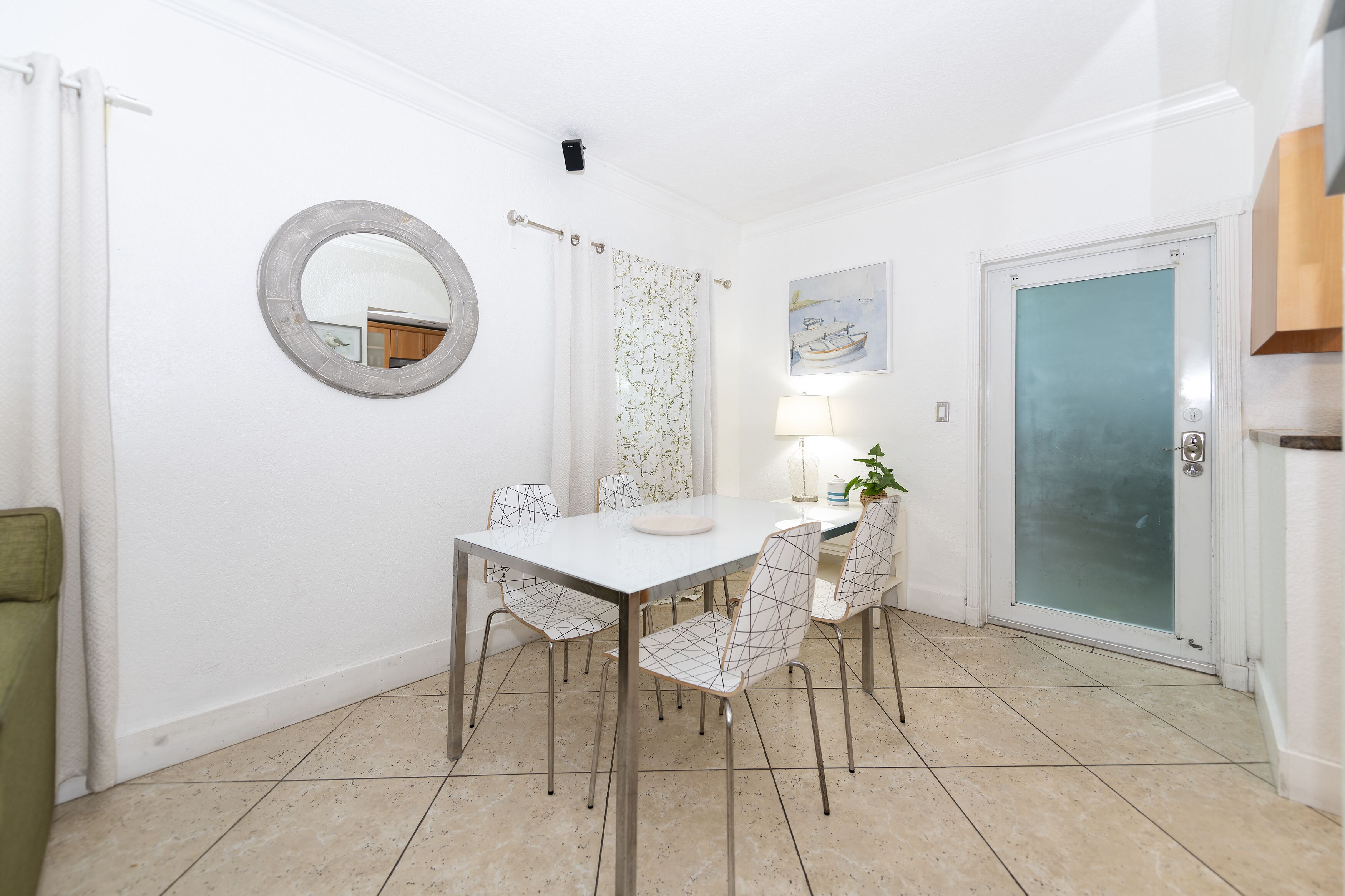 A dining area with a white table, four wireframe chairs, a round mirror, a patterned curtain, a framed picture, and a door with frosted glass.