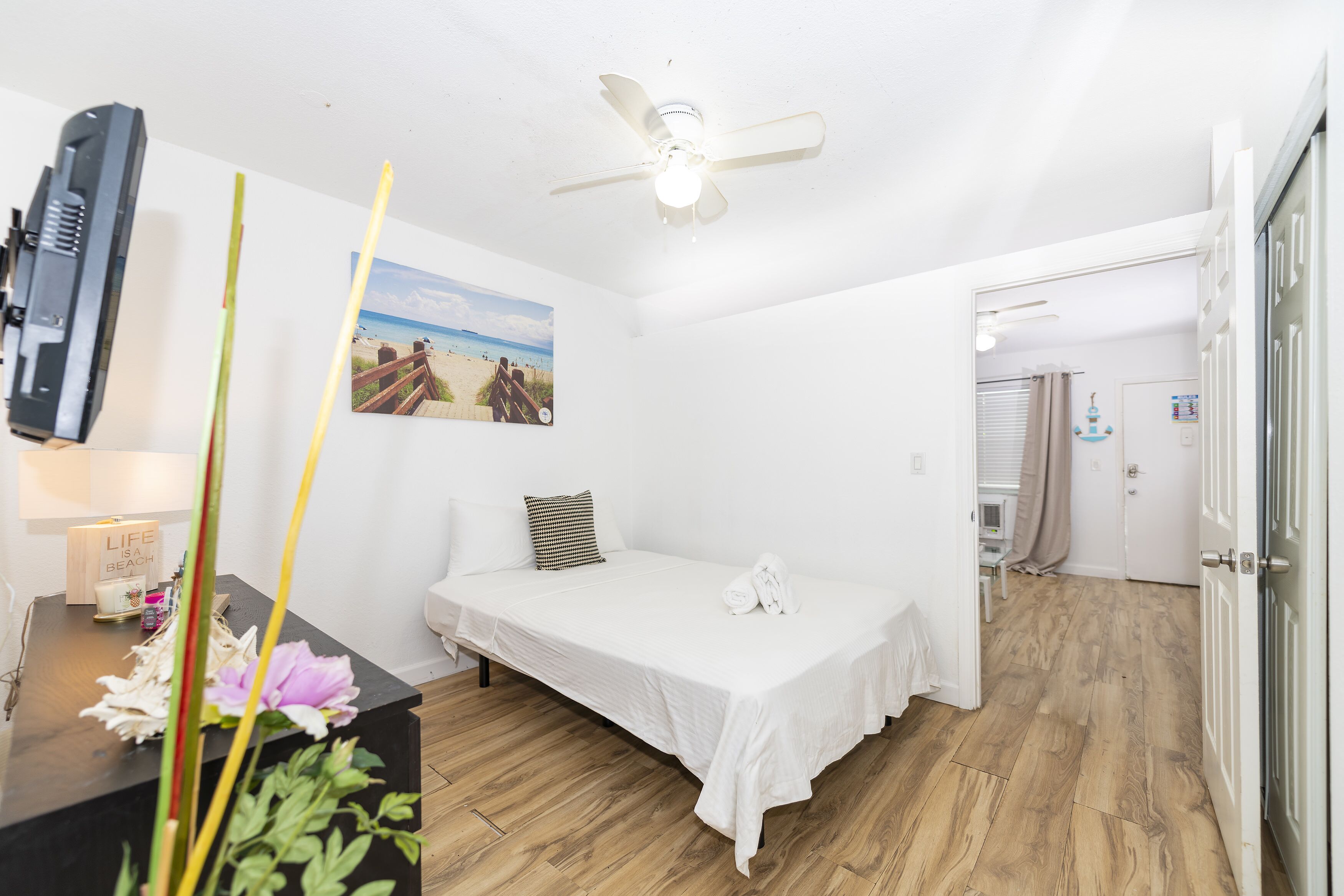 A small, clean room with a single bed, a ceiling fan, a wall-mounted TV, and a beach-themed picture.