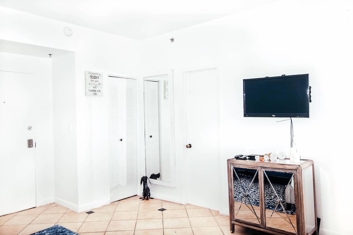 A room with white walls, a tiled floor, a television mounted on the wall, and a wooden cabinet with decorative items.