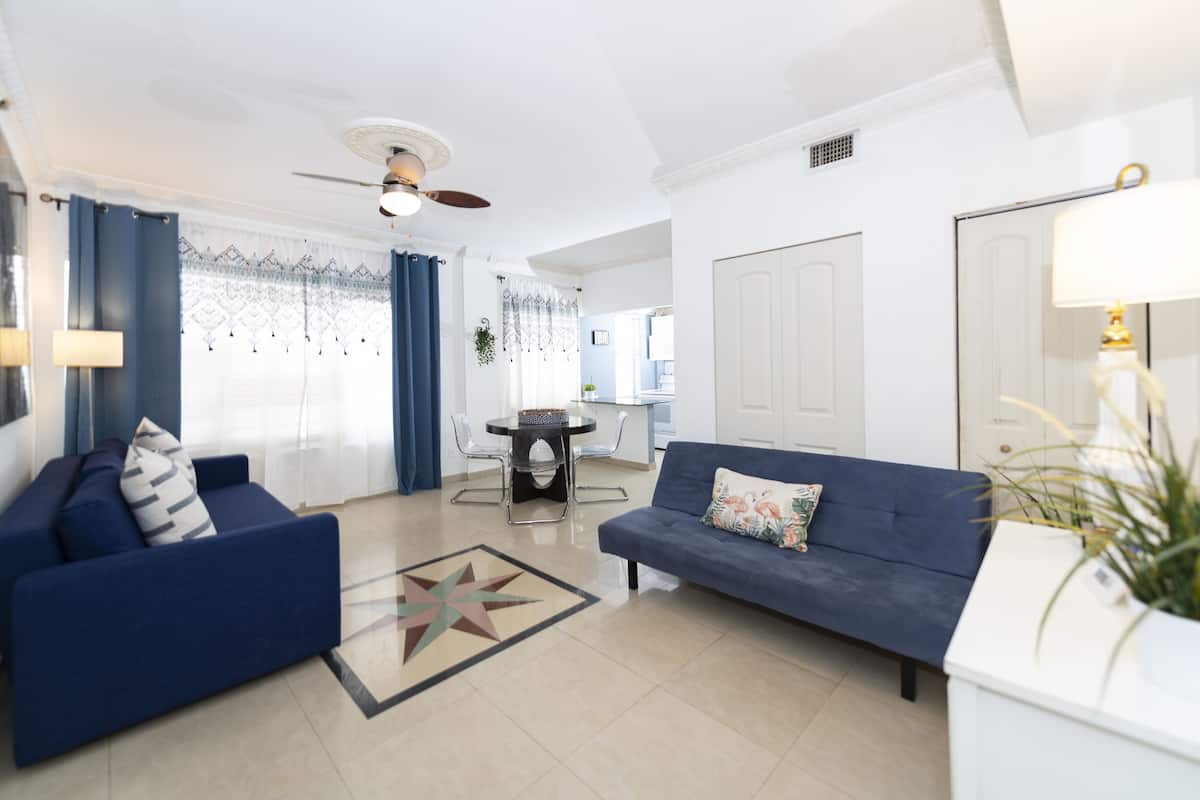 A modern living room with a blue sofa, a matching armchair, a dining table, and a ceiling fan.