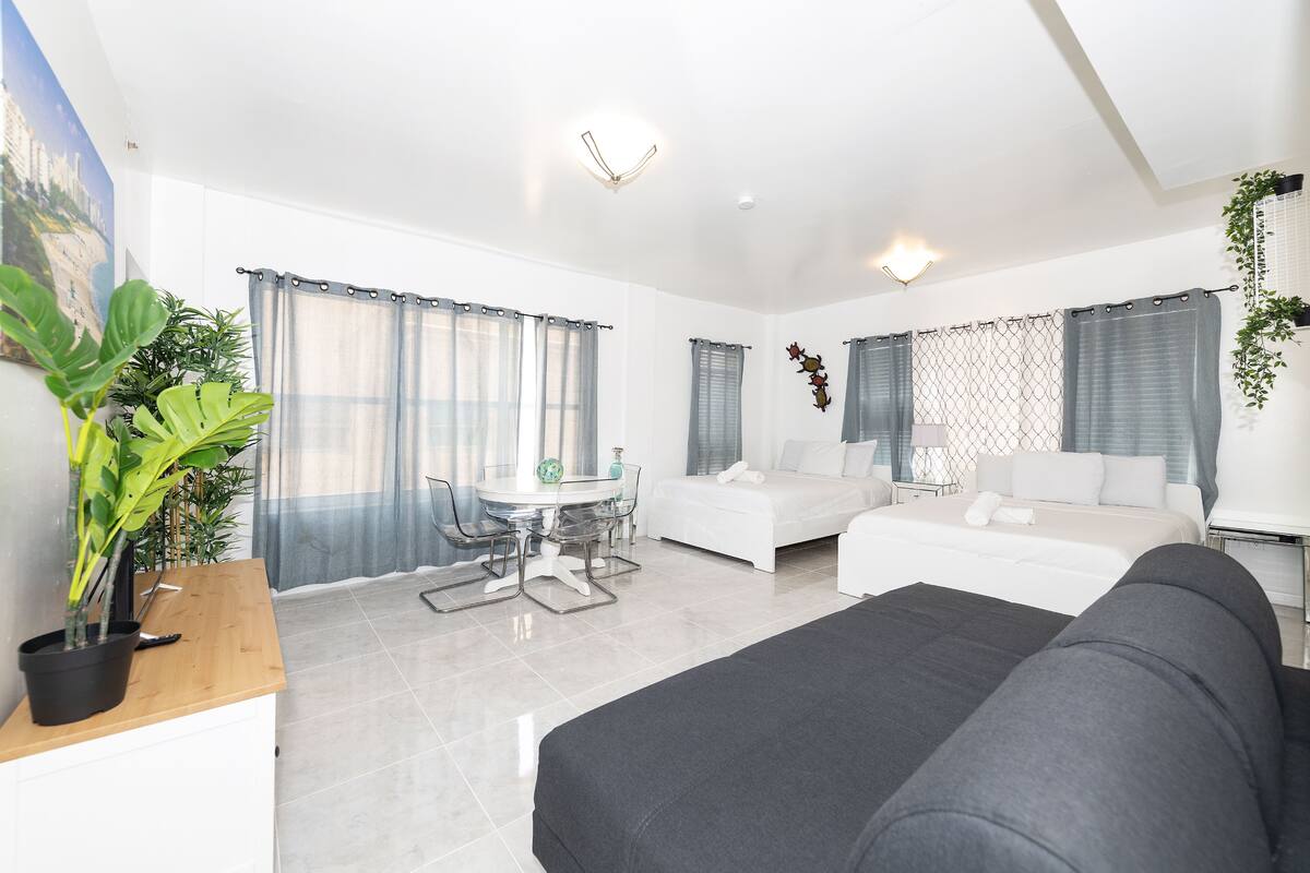 A modern hotel room with a bed, a sofa, a dining table, and a potted plant.