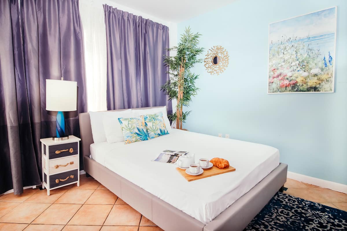 A neatly made bed with a tray of breakfast items, a bedside table with a lamp, and a large window with curtains.