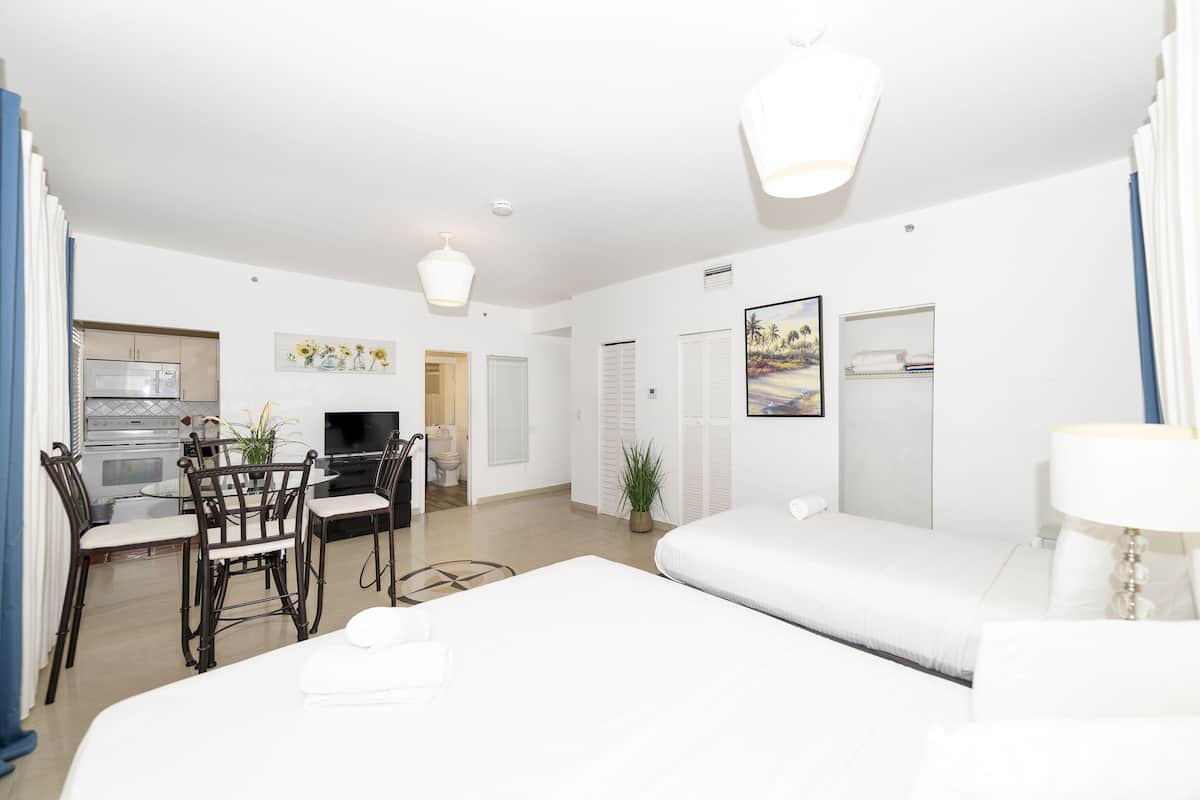 A modern hotel room with a bed, a desk with a chair, a television, and a dining area with chairs and a table.
