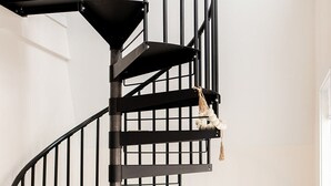 A modern interior with a black spiral staircase, a grey sofa, a wicker chair, and a wooden shelf unit. - Ridge Point | Walk to Glen Arbor | Pet-Friendly | Close to Sleeping Bear Dunes (Glen Arbor)